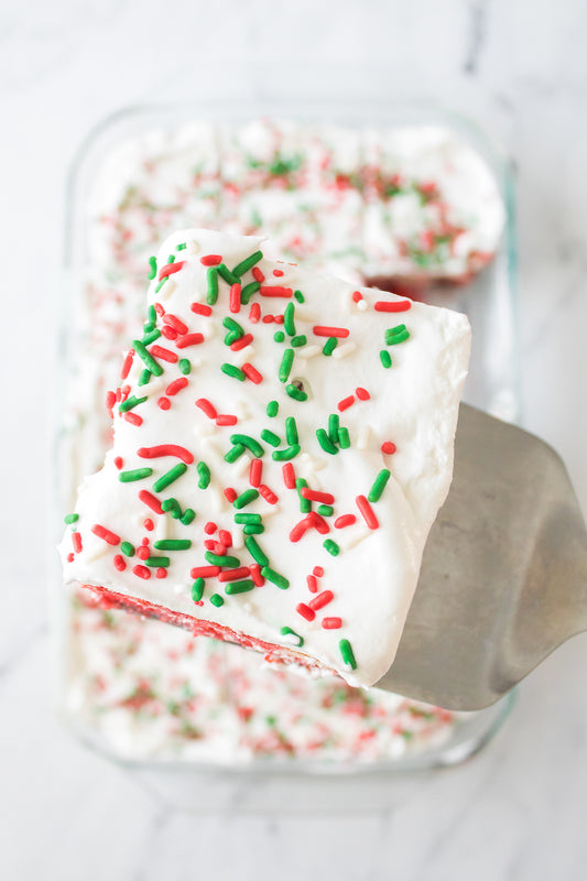 metal spatula with a piece of christmas poke cake topped with cool whip and red, white and green sprinkles